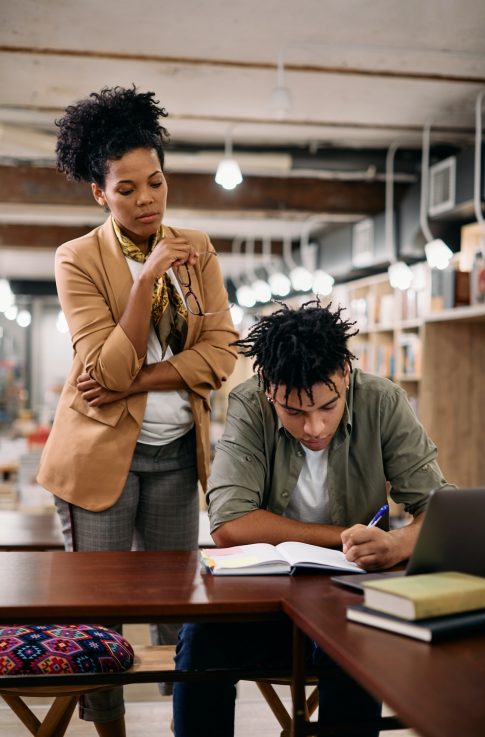 African American professor overseeing her student who is learning in a library.